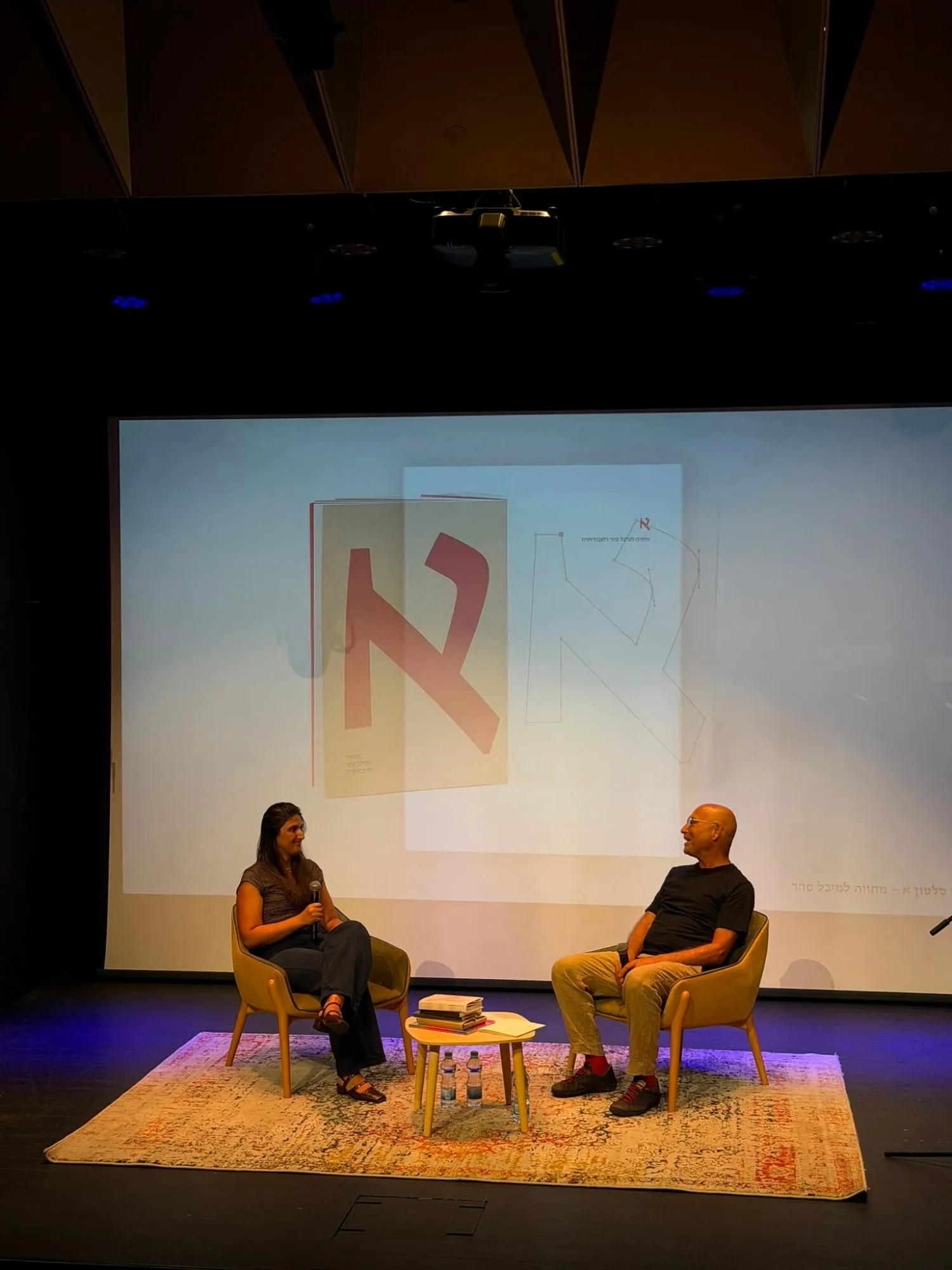 Dana on a stage holding a microphone, a Hebrew letterform project projected on the screen behind her